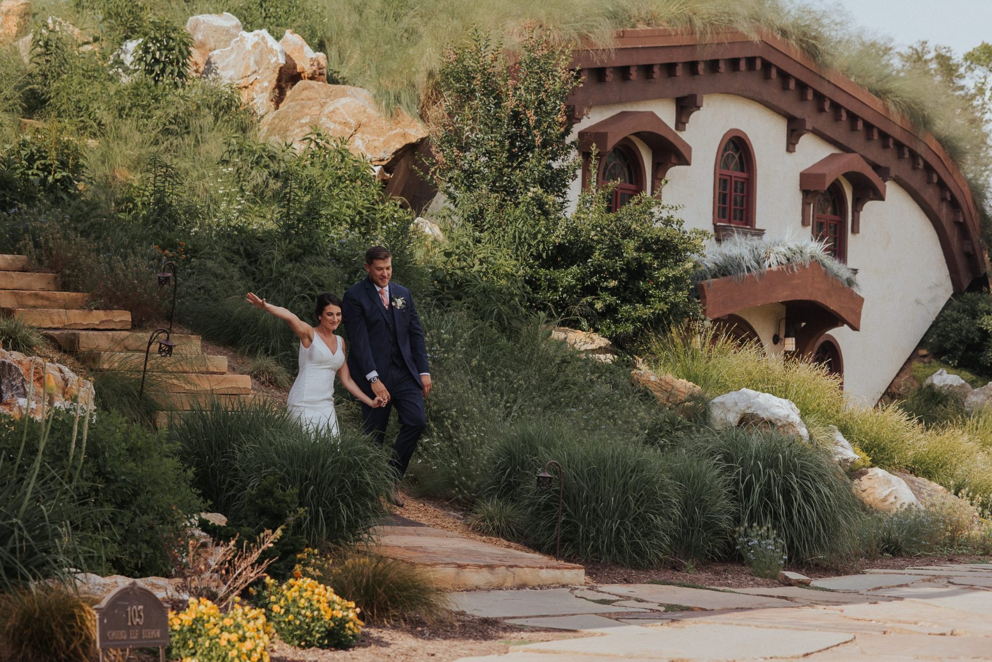 Katie and Luke Wedding A bride and groom walk down stone steps surrounded by greenery, with a whimsical house in the background.