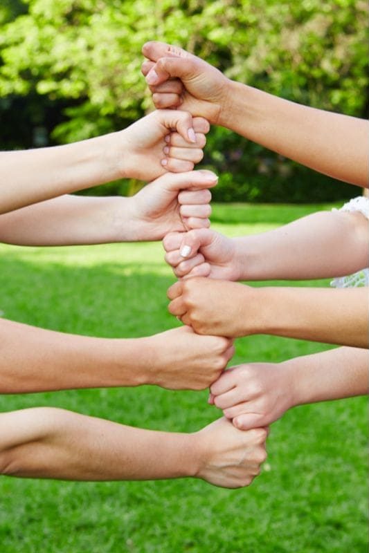 Four pairs of hands grasp each other's wrists in a supportive chain, embodying team empowerment against a backdrop of green grass.