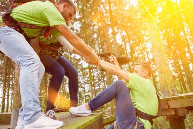 In a sunlit forest, people wearing safety harnesses and green shirts help each other navigate the rope course, showcasing team leadership and empowerment as they plan their next moves together.