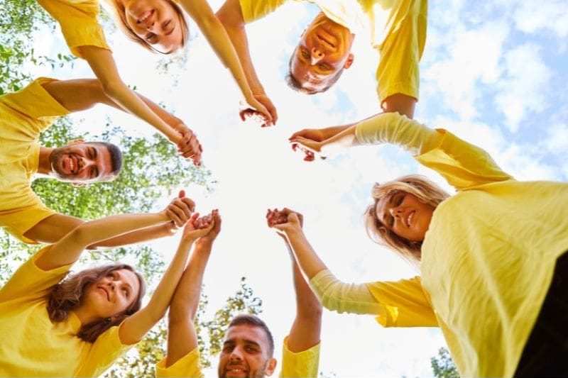 64 A group of people in yellow shirts stand in a circle outdoors, holding hands and looking up at the sky, embodying team leadership and empowerment.