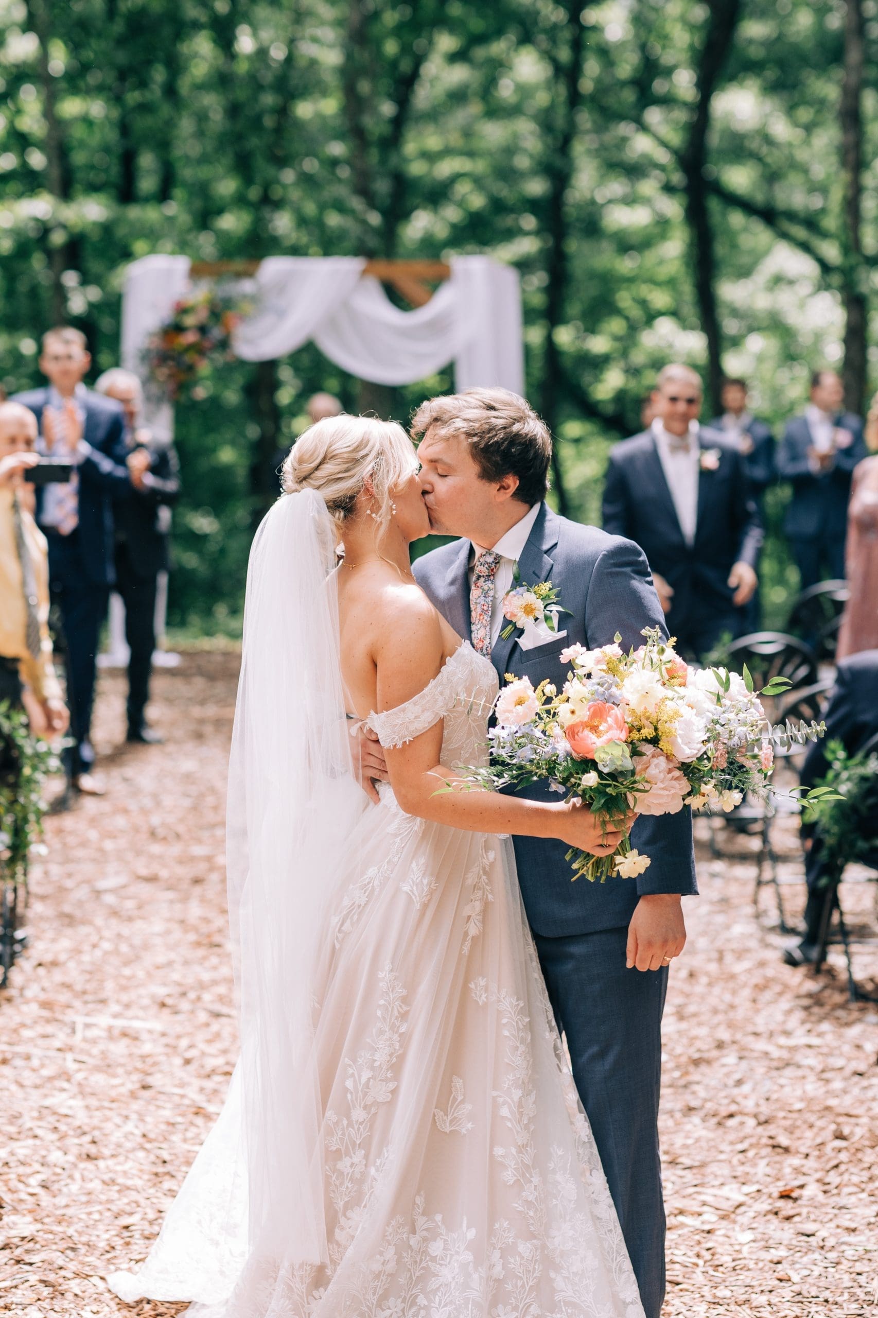 Christie & Connor’s Wedding Day Bride and groom kissing outdoors, surrounded by guests, with a floral arch in the background.
