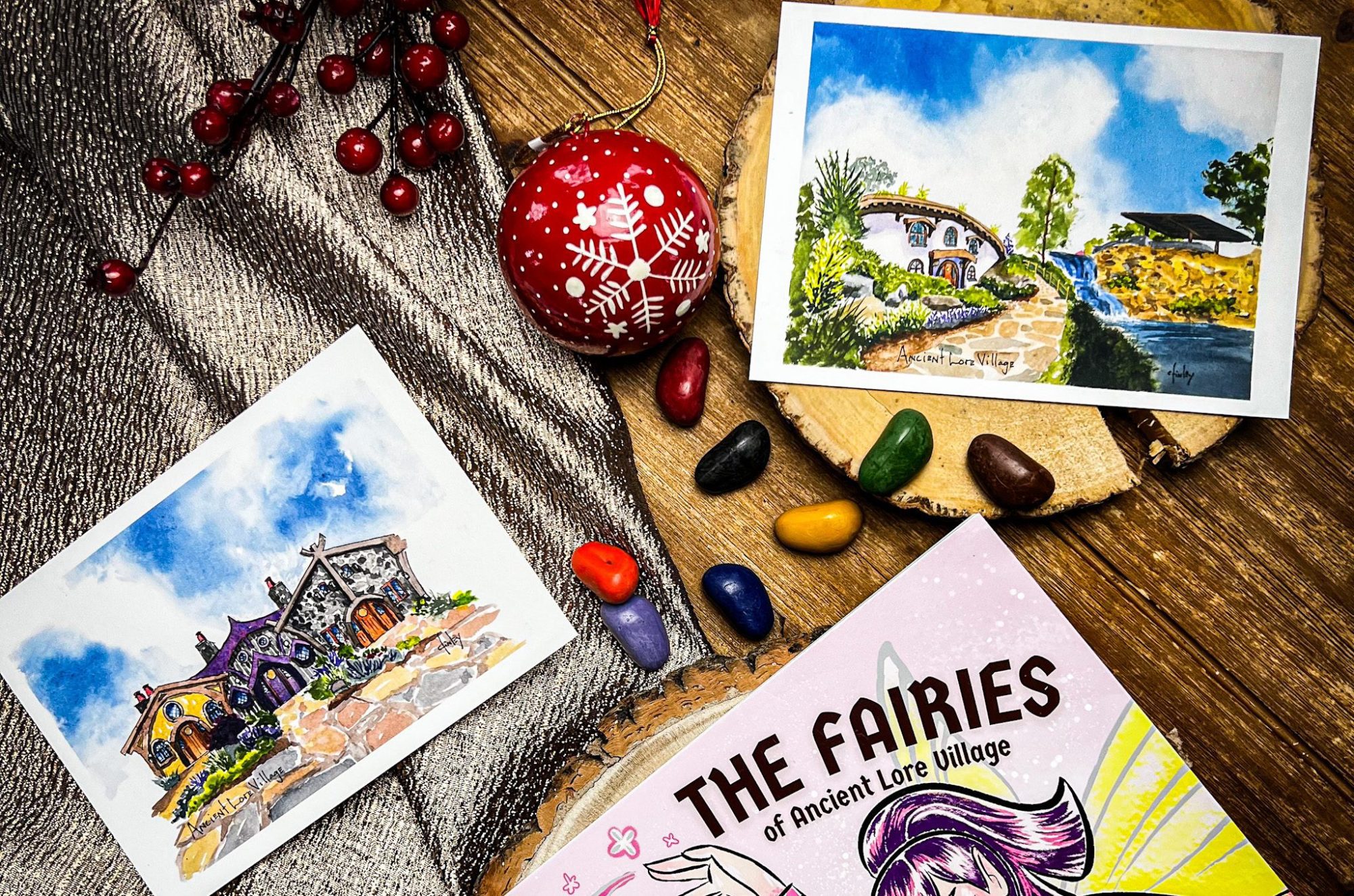 Day 2-Art-1×1 Postcards and a book titled "The Fairies of Ancient Lore Village" are displayed on a wooden surface with some berries and rocks, making an enchanting setup perfect for the gift-giving holidays.