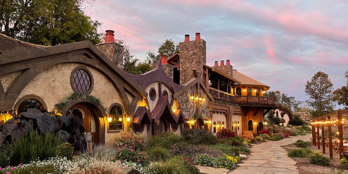 Village Photo Exterior (1200 × 600 px) A whimsical, cottage-style house with rounded doors, lanterns, and lush garden at sunset, resembling a fairy tale setting.