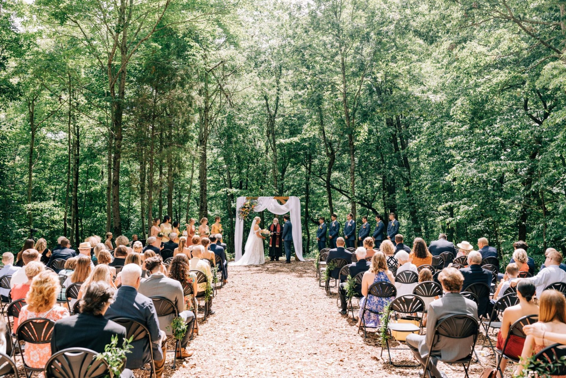 Outdoor wedding in a forest with seated guests watching the couple at the altar under a white arch decorated with flowers.