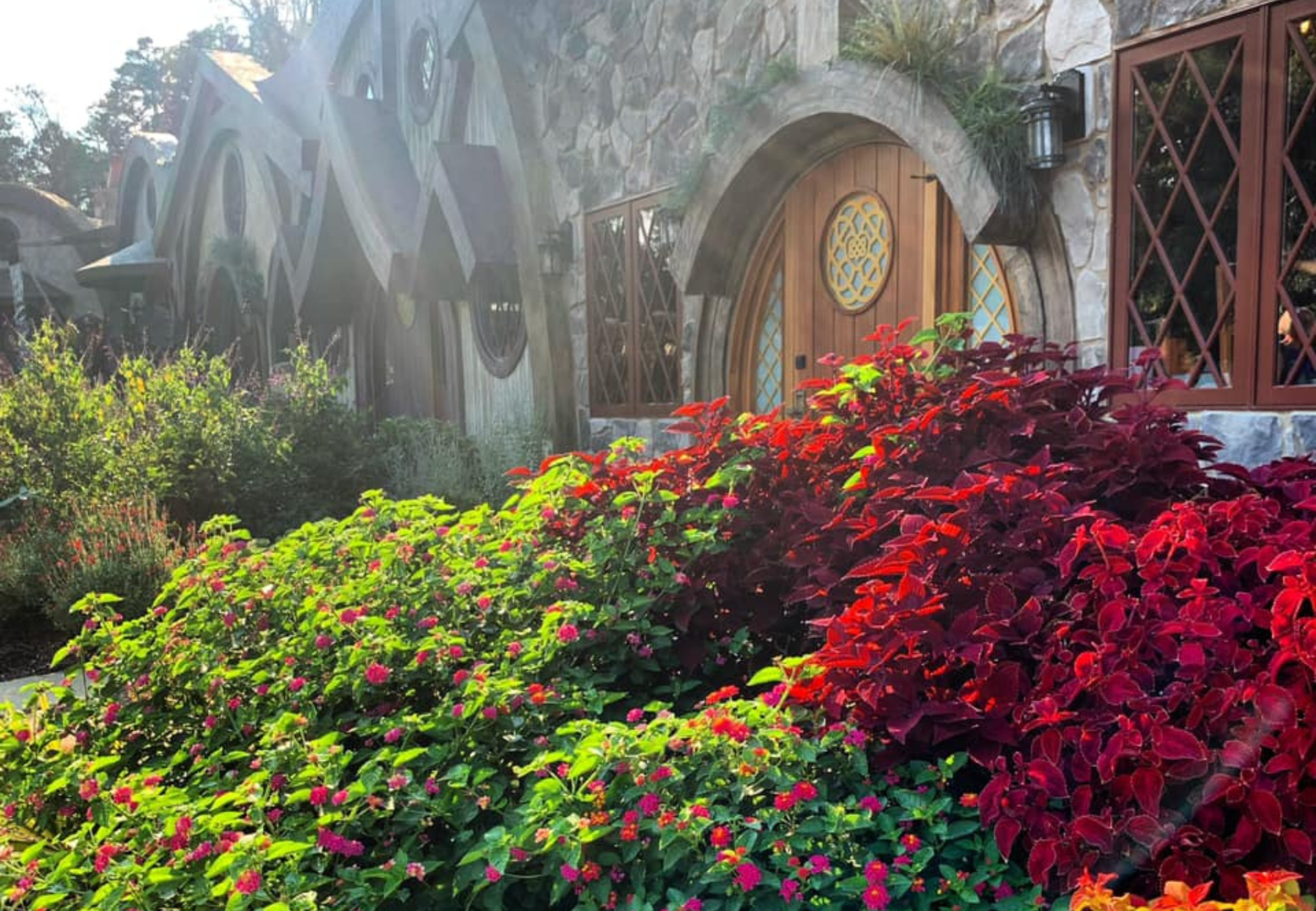 Village Gardens A scenic view of a house with round doors and lush green, red, and pink foliage in the foreground.