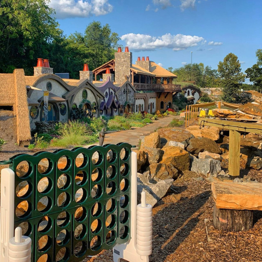 BoardsBottles Outdoor scene with a large Connect Four game in the foreground, whimsical hobbit-like houses in the background, and a clear blue sky. Picture laughter echoing as friends gather around, enjoying charcuterie boards and wine while playing yard games under the sun.
