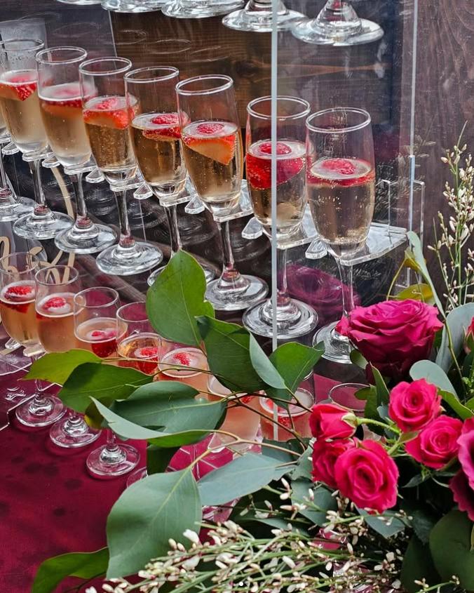 Rows of champagne glasses with raspberries, accompanied by a bouquet of pink and red flowers on a burgundy cloth.